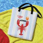 A striped tote bag with black handles sits on a yellow towel near a pool, featuring a red lobster and the text 'GÉZUAR!' on the front.