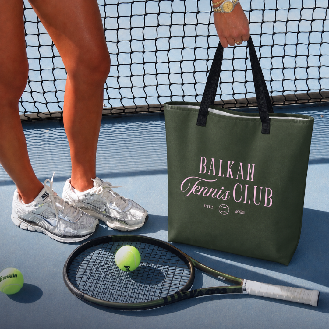 An olive green canvas tote bag with black handles sits on a blue tennis court next to a player's feet, a tennis racket, and tennis balls. The bag features the text 'BALKAN Tennis CLUB' in pink and white.