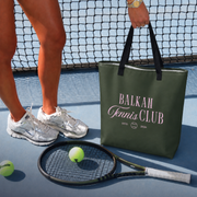 An olive green canvas tote bag with black handles sits on a blue tennis court next to a player's feet, a tennis racket, and tennis balls. The bag features the text 'BALKAN Tennis CLUB' in pink and white.