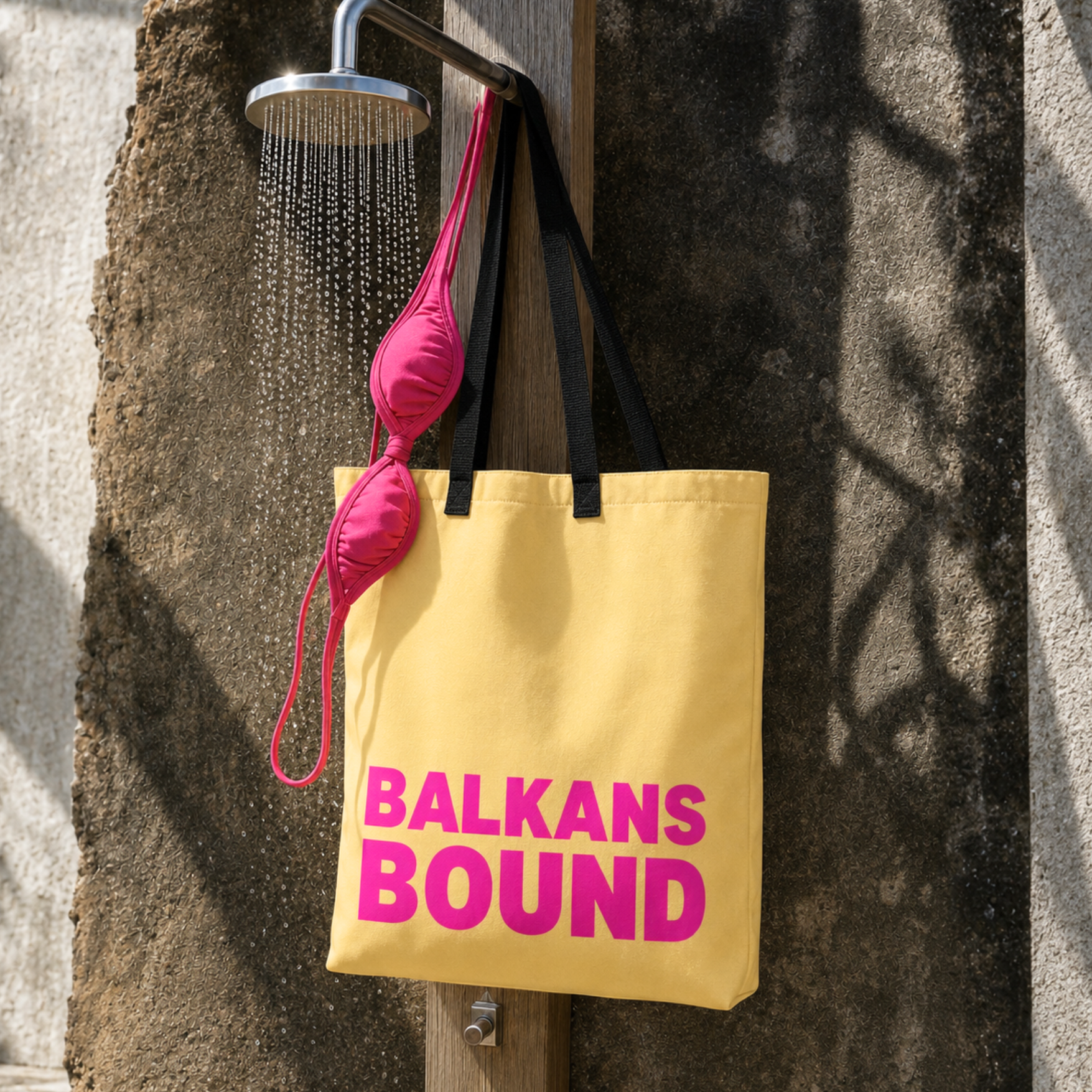 A yellow canvas tote bag with black handles and bold pink text reading 'BALKANS BOUND' hangs from a shower rod under a shower head, with a pink bikini top draped over the bag against a textured concrete wall.