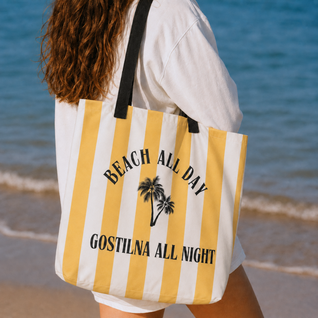 A person at the beach carrying a large white tote bag with yellow vertical stripes, black straps, and black text reading 'BEACH ALL DAY' and 'GOSTILNA ALL NIGHT' with a small palm tree graphic.