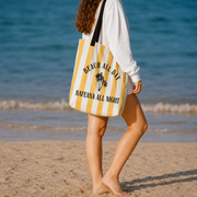 A woman on a sandy beach walks barefoot while carrying a large yellow and white striped tote bag with black text that reads 'BEACH ALL DAY KAFEANA ALL NIGHT' and a small palm tree graphic.