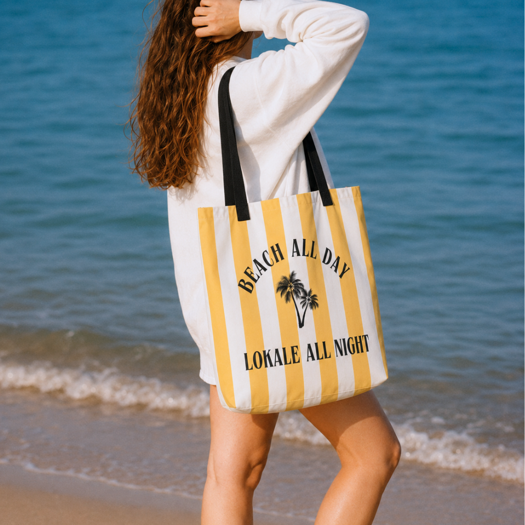 A woman on a beach carries a large striped tote bag with yellow and white vertical stripes, black handles, and the text 'BEACH ALL DAY' and 'LOKALE ALL NIGHT' with a palm tree graphic.