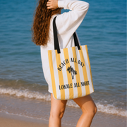 A woman on a beach carries a large striped tote bag with yellow and white vertical stripes, black handles, and the text 'BEACH ALL DAY' and 'LOKALE ALL NIGHT' with a palm tree graphic.