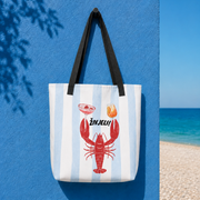 A white canvas tote bag with light blue vertical stripes, featuring a red lobster graphic, the word 'Živjeli' in black, and two black shoulder straps. The bag is hanging against a bright blue wall next to a beach scene with the ocean and sand visible.