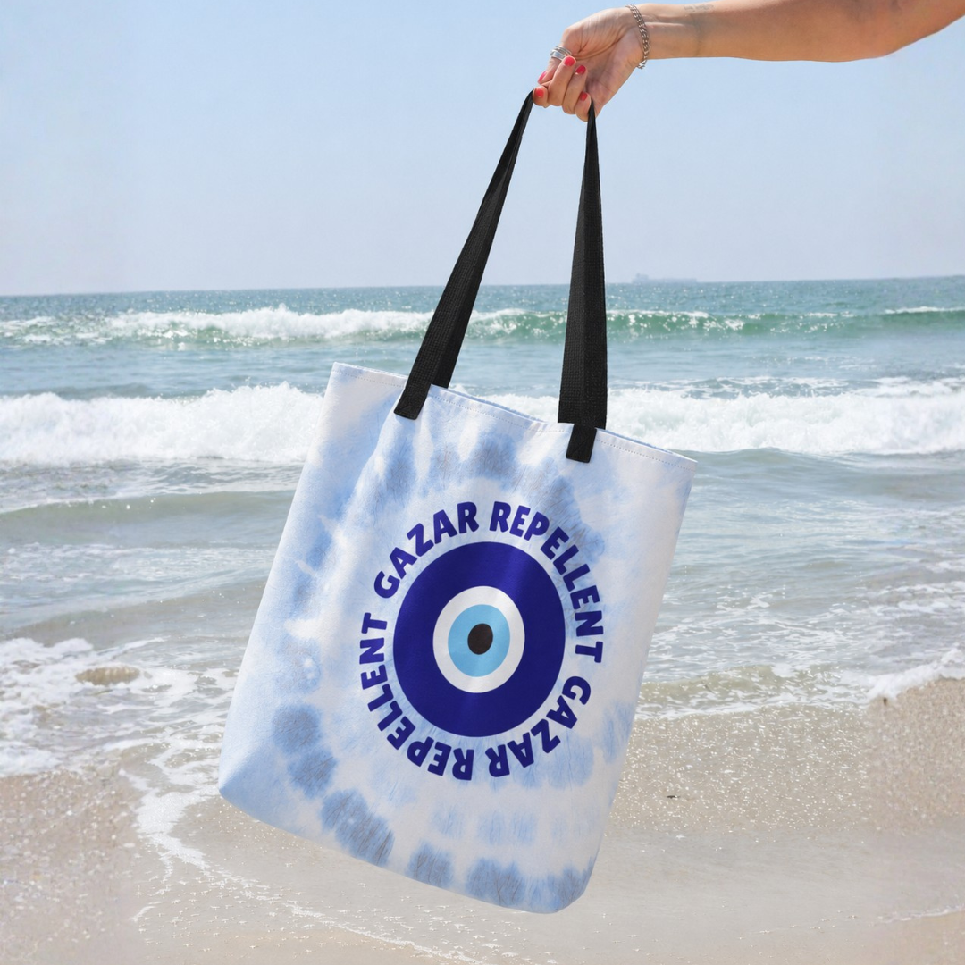 A white and light blue tie-dye beach tote bag with black handles, held by a hand against a beach background. The bag features a large blue evil eye design with the text 'GAZAR REPELLENT'.