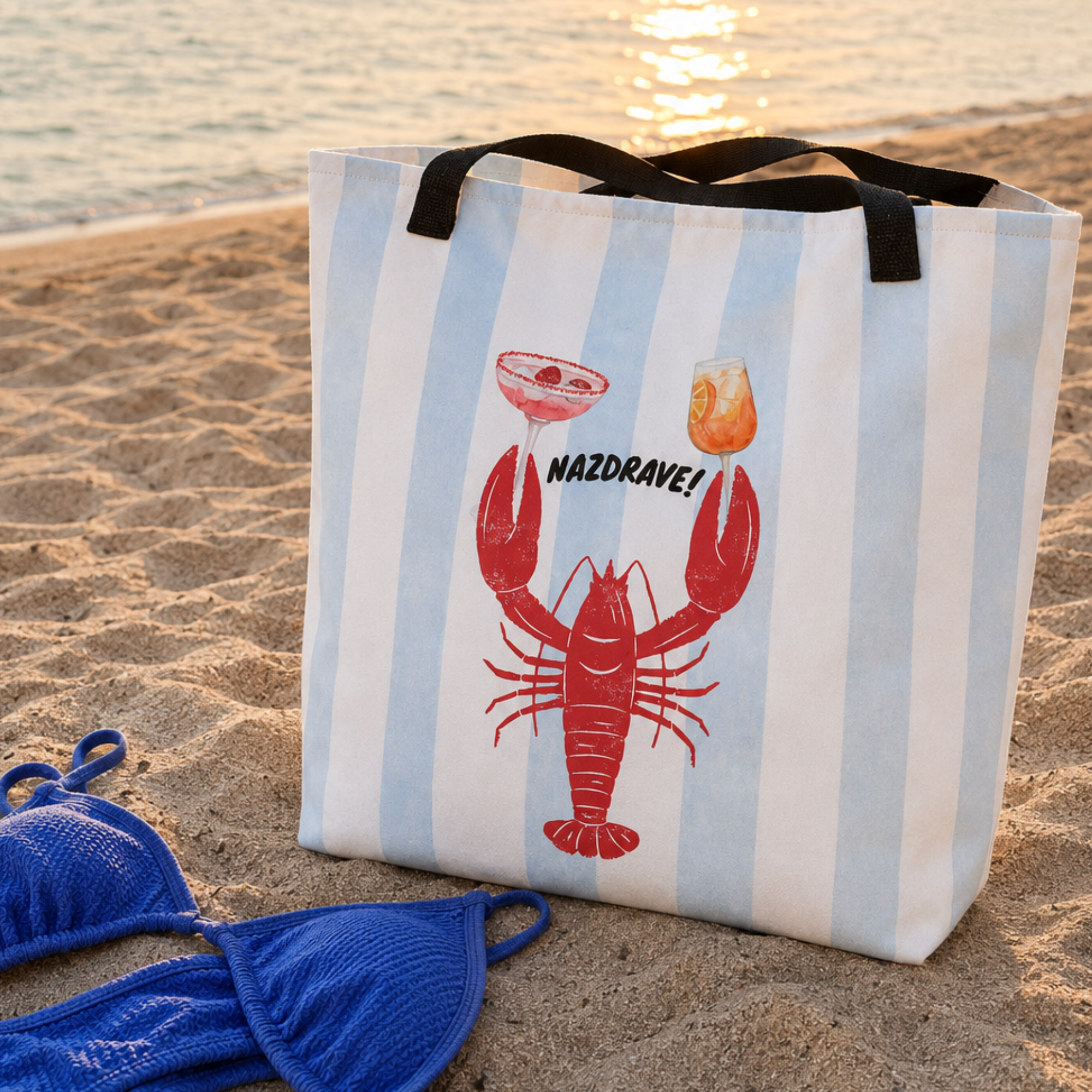 A beach tote bag with light blue and white vertical stripes stands on a sandy beach. The bag features a red lobster graphic, a cocktail glass with the word 'NAZDRAVE!', and black handles.