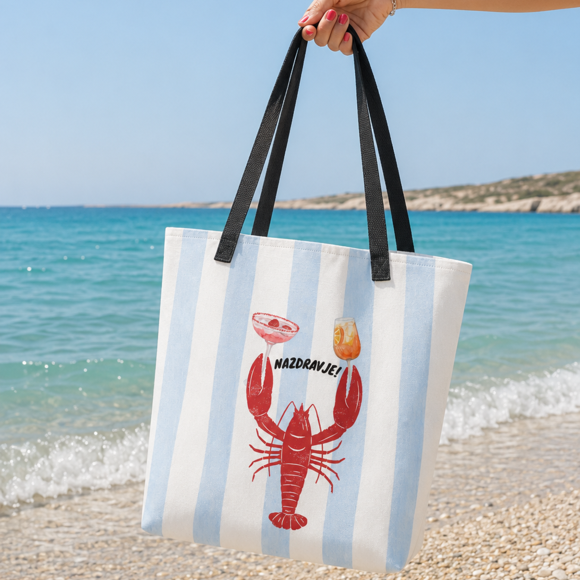 A white canvas beach tote bag with light blue vertical stripes, black shoulder straps, and a red lobster graphic with a cocktail and a small orange item, held by a hand against a beach and ocean background.