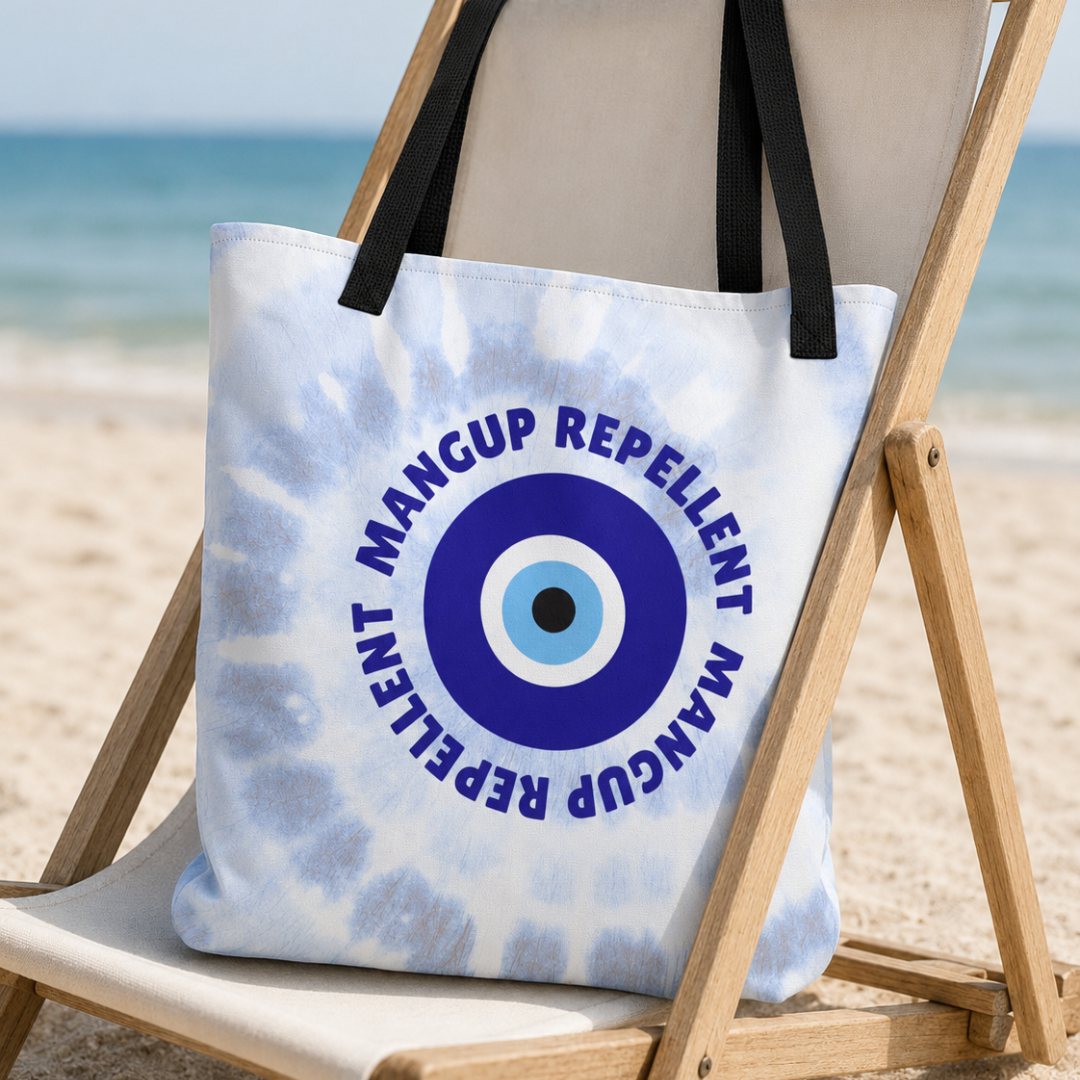 A white and light blue tie-dye tote bag with black handles sits on a wooden beach chair at the beach. The bag features a circular evil eye graphic in blue, white, and black, surrounded by the text 'MANGUP REPELLENT'.