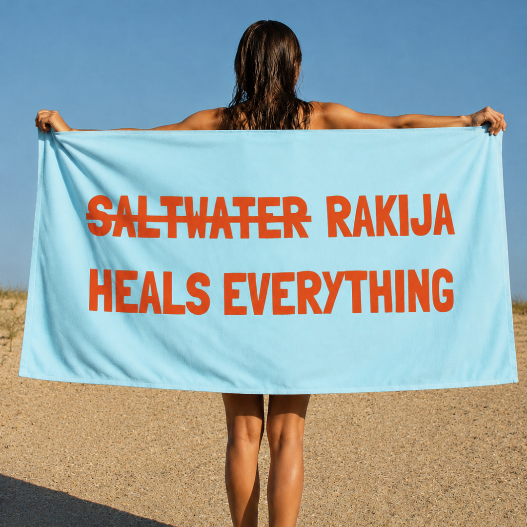 A woman stands on a sandy beach holding up a large light blue towel or beach wrap with bold orange text that reads 'SALTWATER RAKIJA HEALS EVERYTHING' against a clear blue sky.