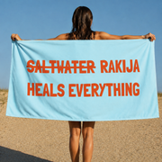 A woman stands on a sandy beach holding up a large light blue towel or beach wrap with bold orange text that reads 'SALTWATER RAKIJA HEALS EVERYTHING' against a clear blue sky.