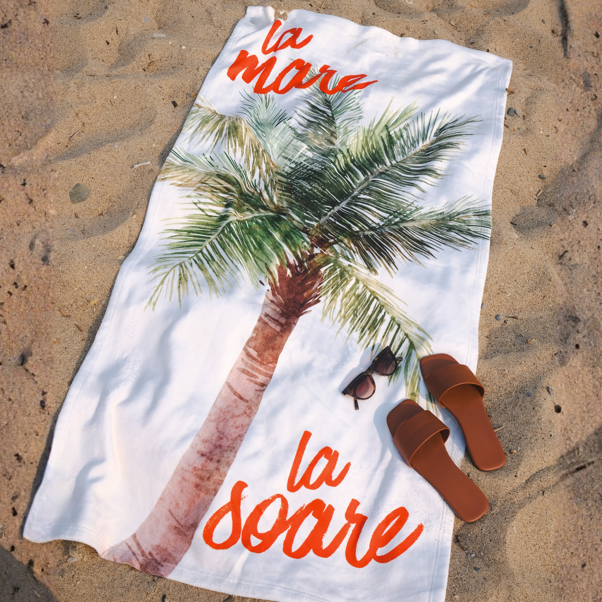 A white beach towel laid flat on sandy ground, printed with a large palm tree and the orange script text 'la mare la soare'. Brown slide sandals and a pair of sunglasses are placed on the towel as styling props.