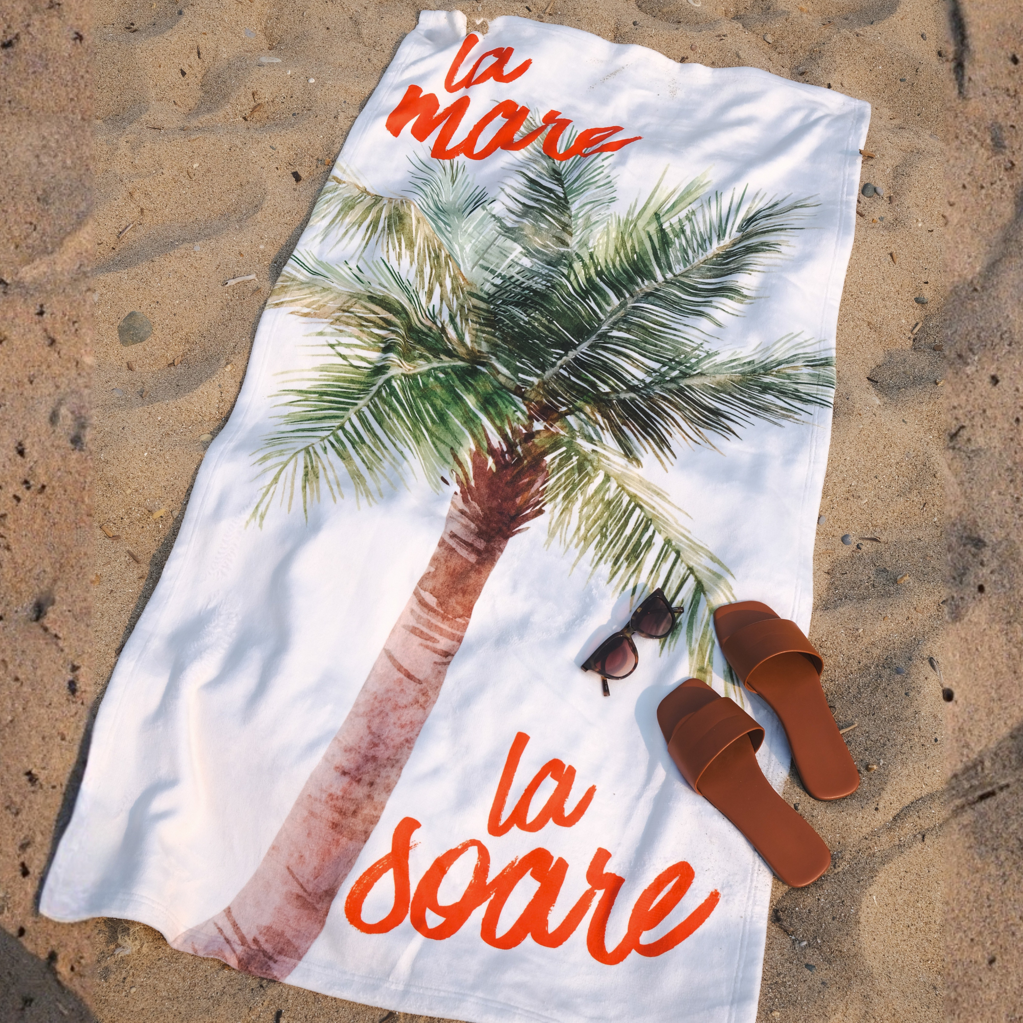 A white beach towel laid flat on sandy ground, printed with a large palm tree and the orange script text 'la mare la soare'. Brown slide sandals and a pair of sunglasses are placed on the towel as styling props.