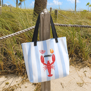 A canvas tote bag with light blue and white vertical stripes, featuring a red lobster graphic and the word 'NOROC!' on the front, hanging from a wooden post on a sandy beach path with grass in the background.