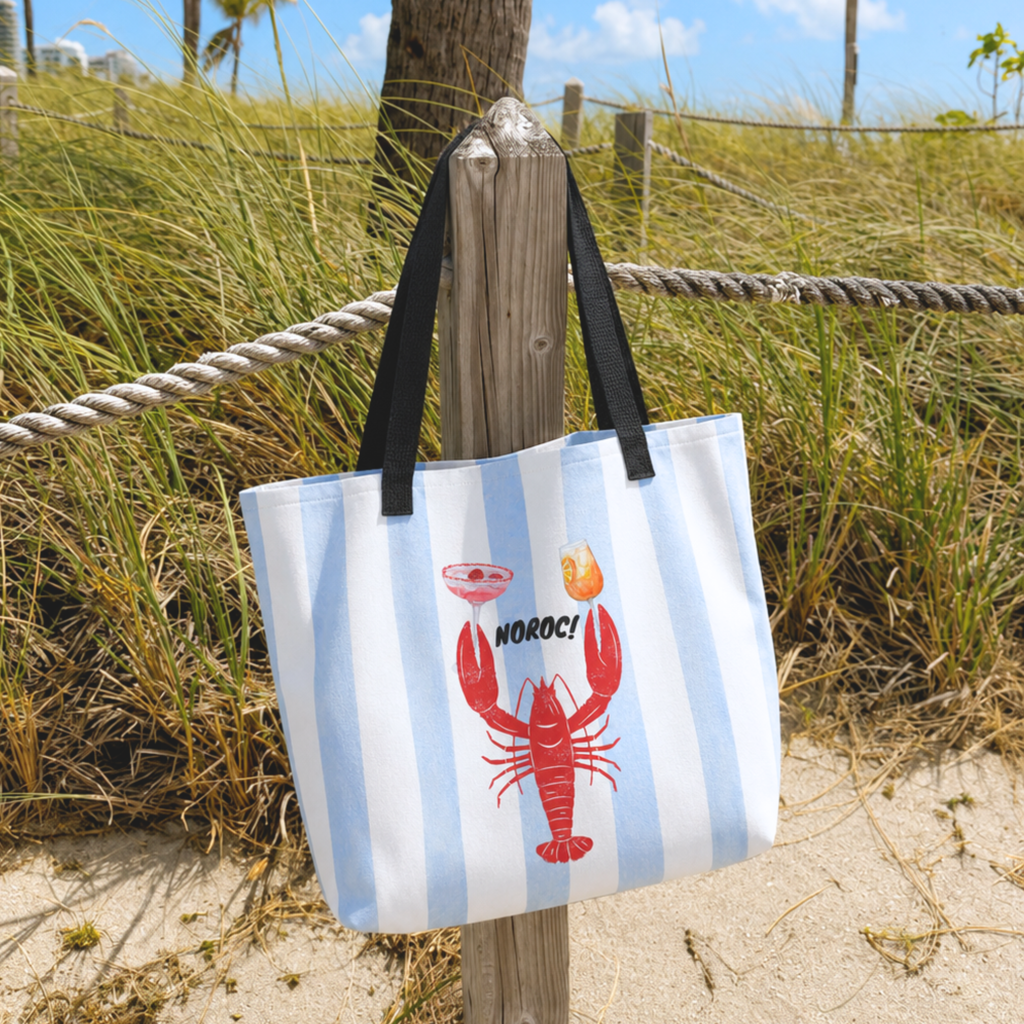 A canvas tote bag with light blue and white vertical stripes, featuring a red lobster graphic and the word 'NOROC!' on the front, hanging from a wooden post on a sandy beach path with grass in the background.