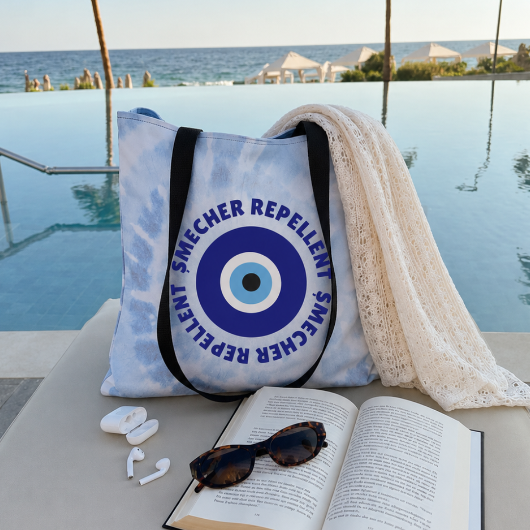 A light blue and white tie-dye tote bag with black handles and a large blue circular emblem reading “SMECHER REPELLENT,” placed by a pool with an open book, sunglasses, earbuds, and a cream lace throw nearby.