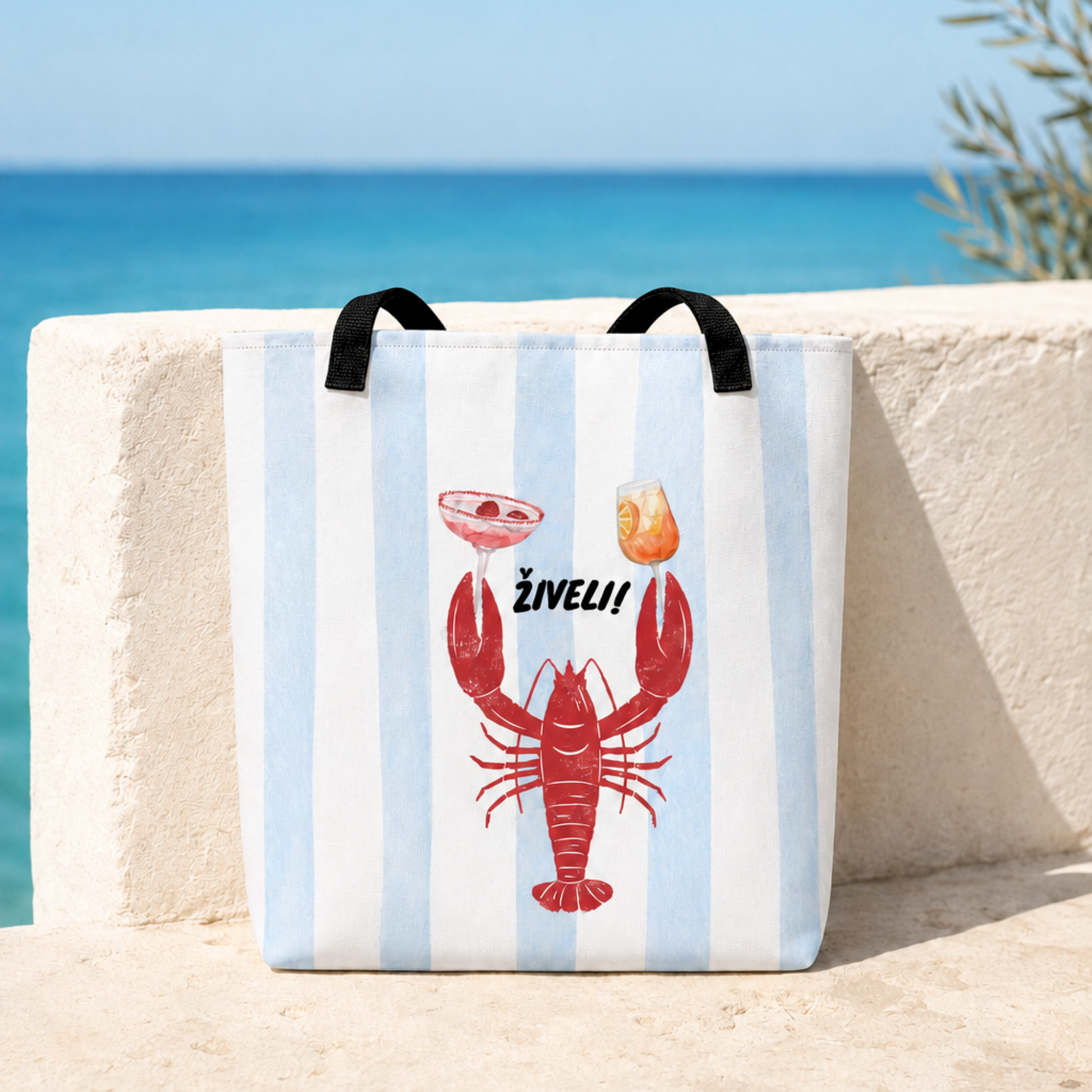 A canvas tote bag with light blue and white vertical stripes, featuring a red lobster graphic holding a cocktail and an orange drink, with the text 'żIVELI!' above it. The bag has black handles and is photographed outdoors leaning against a light-colored stone wall with a blue ocean background.