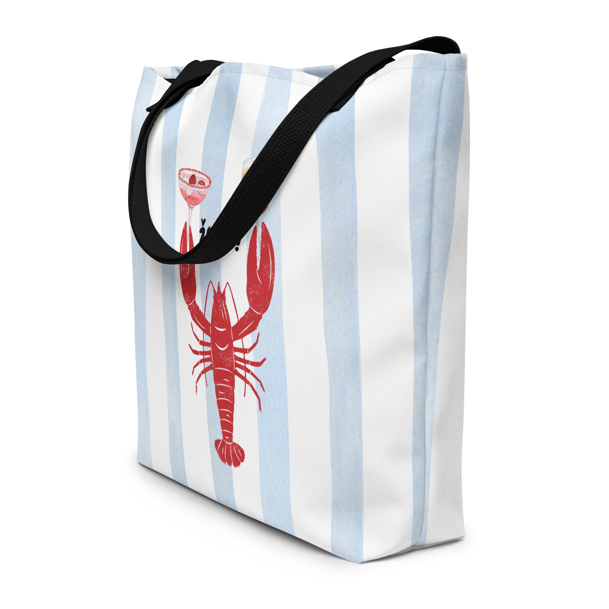 A canvas tote bag with light blue and white vertical stripes, featuring a red lobster graphic holding a cocktail and an orange drink, with the text 'żIVELI!' above it. The bag has black handles and is photographed outdoors leaning against a light-colored stone wall with a blue ocean background.