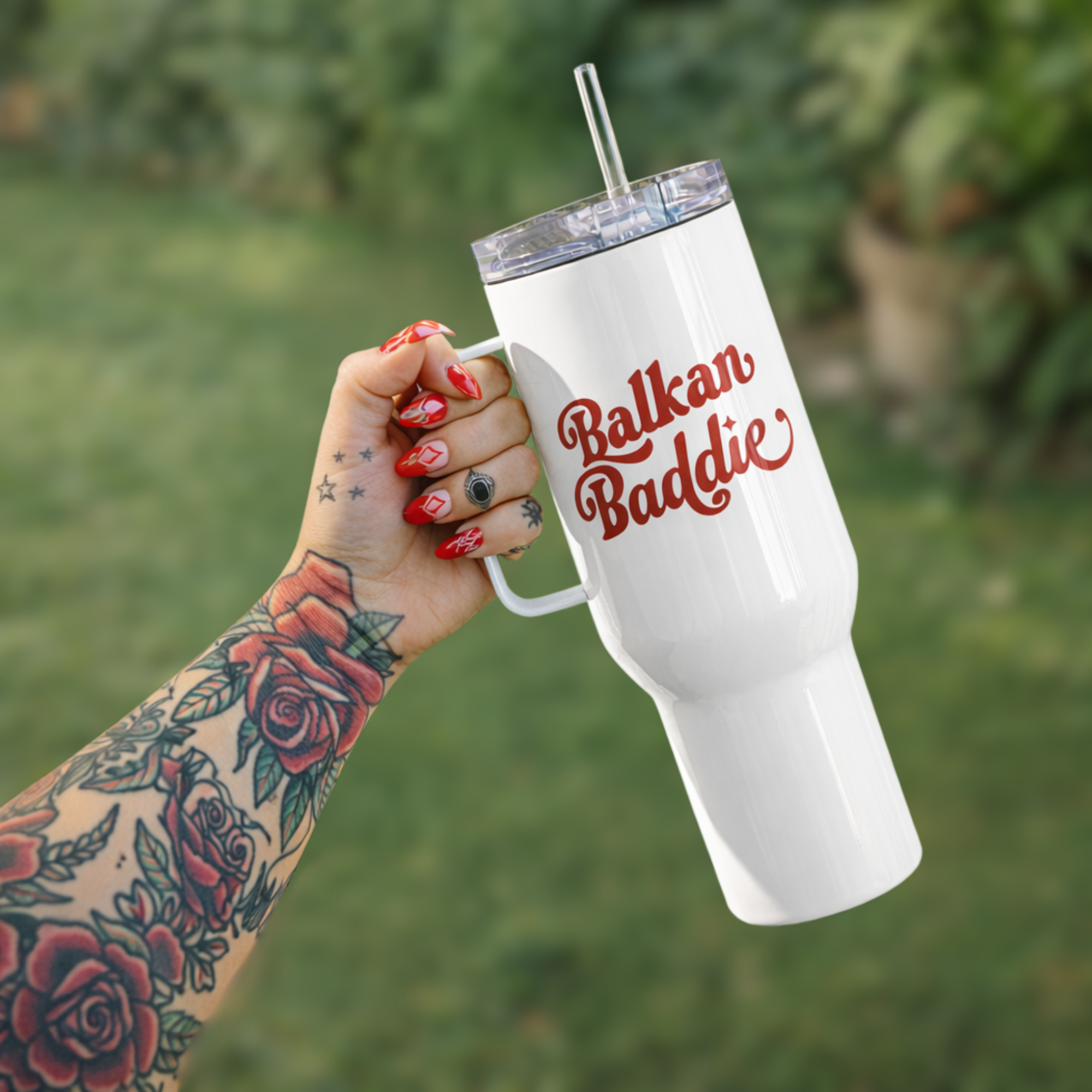 White tumbler with 'Balkan Baddie' text held by a tattooed hand against a blurred green background