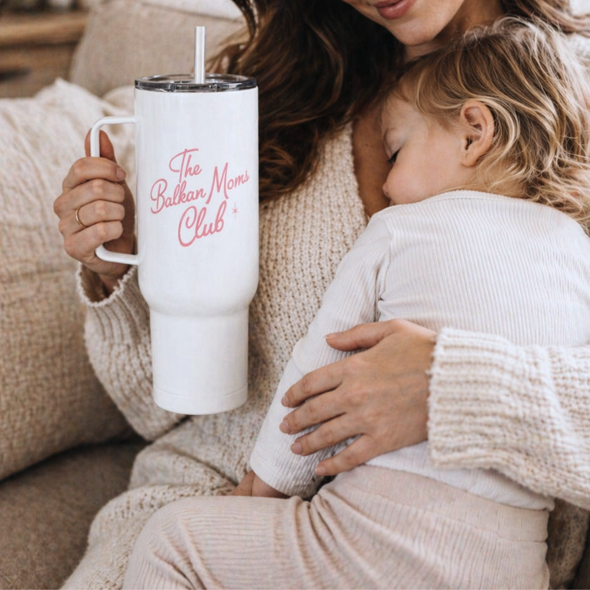 Woman holding a white tumbler with 'The Balkan Moms Club' text, sitting on a couch with a child.