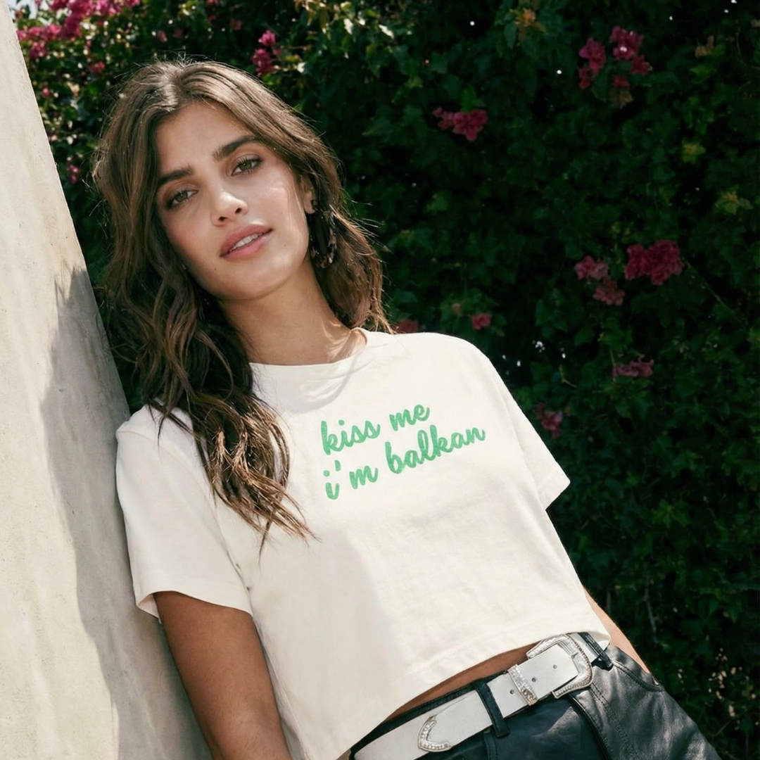 Model wearing white short-sleeved crop top that says "Kiss me I'm Balkan" in green lettering outdoors leaning against a building