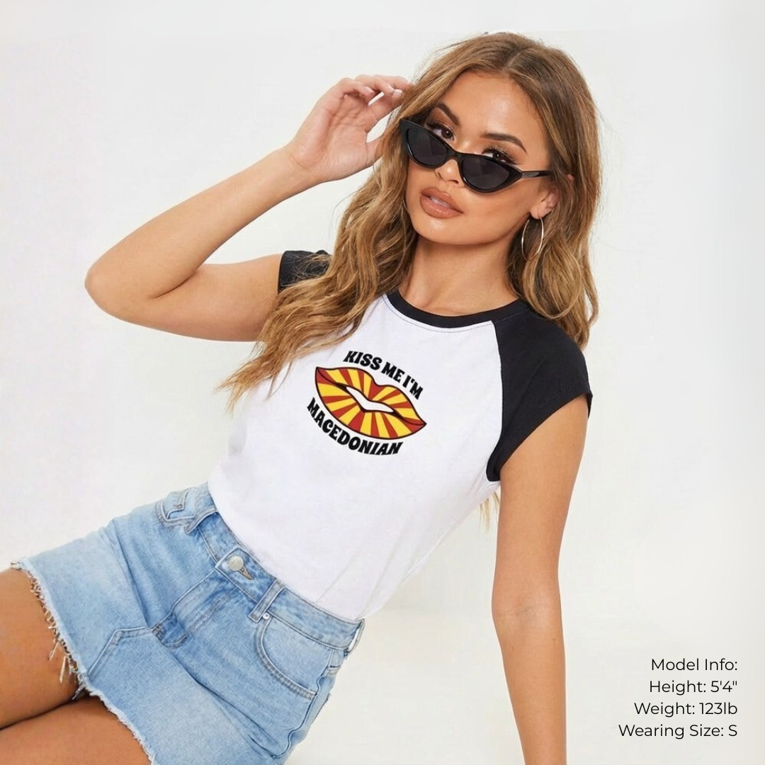 Model sitting on the ground on white studio backdrop wearing a mini denim shirt, black cat-eye sunglasses and a white short sleeved t-shirt with black sleeves an red and white striped lips on the the t-shirt that says "Kiss Me I'm Macedonian"