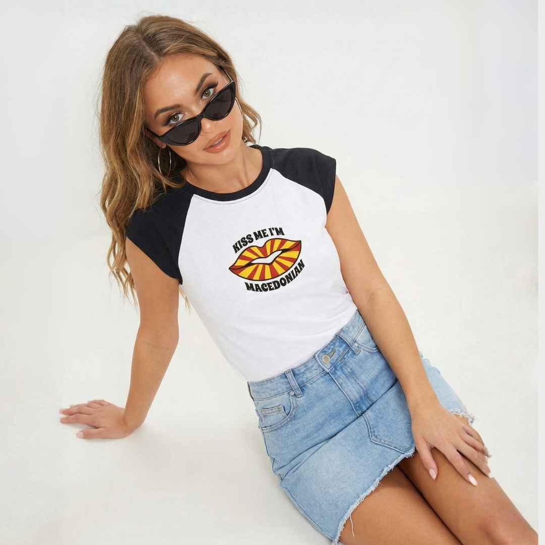 Model sitting on the ground on white studio backdrop wearing a mini denim shirt, black cat-eye sunglasses and a white short sleeved t-shirt with black sleeves an red and white striped lips on the the t-shirt that says "Kiss Me I'm Macedonian"