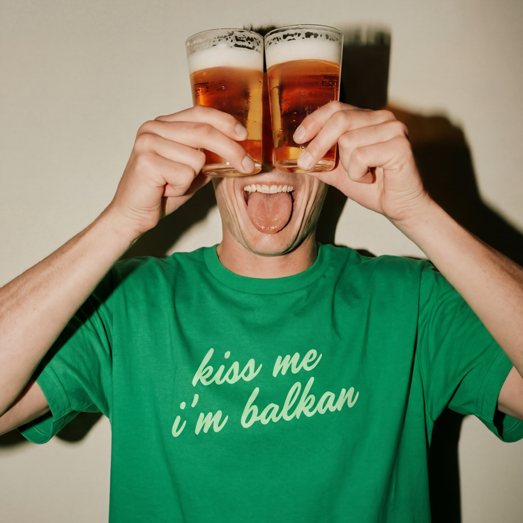 Person wearing a green t-shirt with 'kiss me i'm balkan' text, holding two glasses of beer.