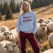 A woman stands in a grassy mountain meadow with sheep around her, wearing a light blue sweatshirt with red script text reading 'Mentally in the Balkans' and burgundy track pants with white side stripes.